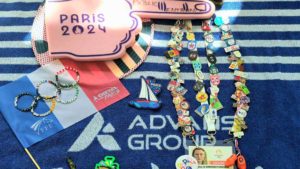 Collection de pin's récoltés lors des Jeux Olympiques de Paris 2024, accompagnée de souvenirs tels qu'un drapeau français, un badge, et une main en mousse, disposés sur une serviette aux couleurs d'Advans Group.