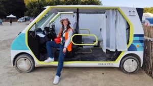 Amandine aux Jeux Olympiques 2024, posant souriante devant un véhicule autonome utilisé sur le site, équipée de son gilet orange.